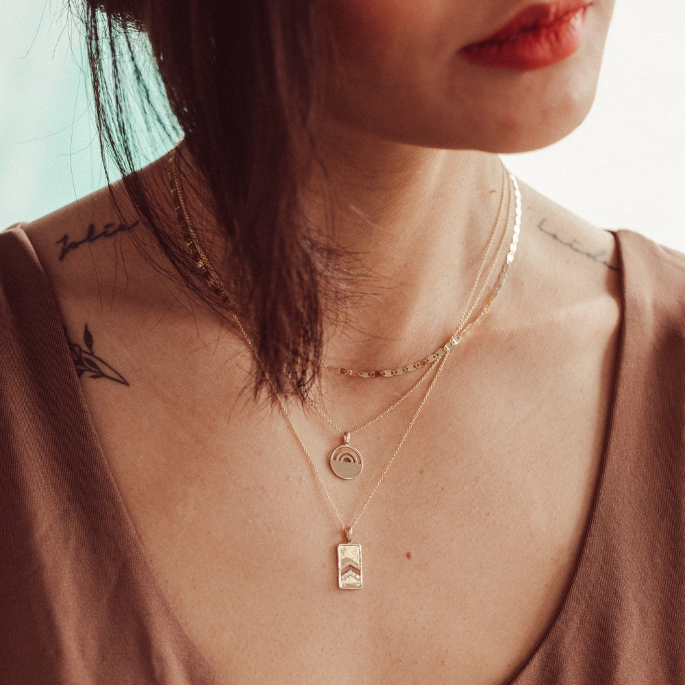 Close-up of a person wearing a  gold necklace with a pendant, set against a blurred background.