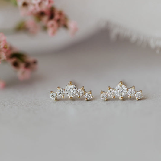 A pair of tapered curved diamond stud earrings in 14k gold, displayed against a soft background with floral elements.