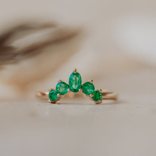 A gold band ring with multiple round and oval emeralds set in a starburst design, placed on a neutral background.