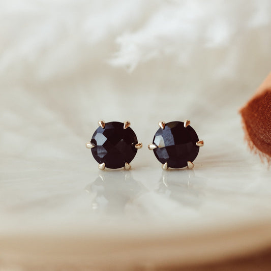 A pair of 14k gold stud earrings featuring round black spinel stones set in six prongs, displayed on a white platform with a textured background.