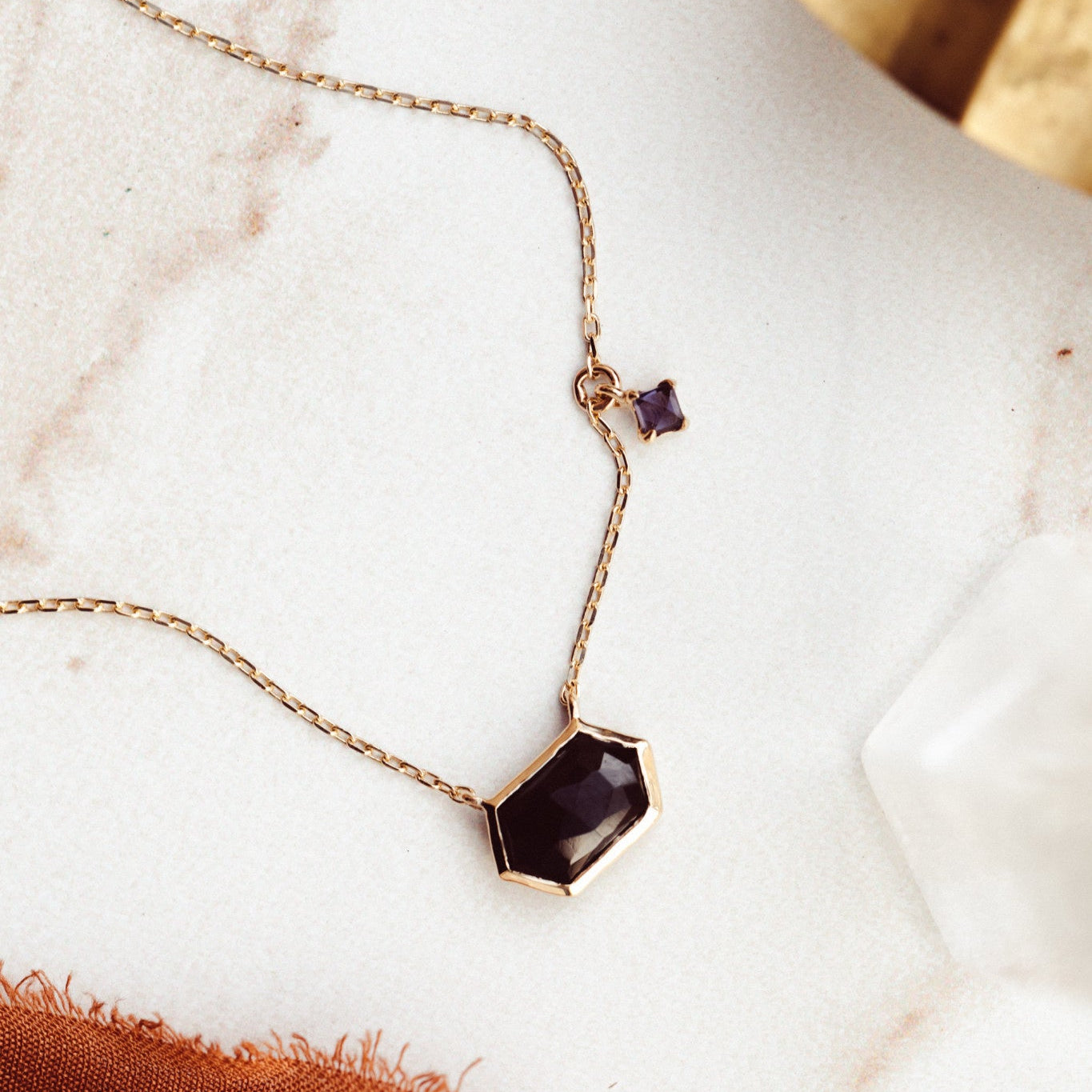 A long hexagon black spinel necklace with a small square iolite dangle, set in a high polish bezel, displayed against a marble background with a gold chain.