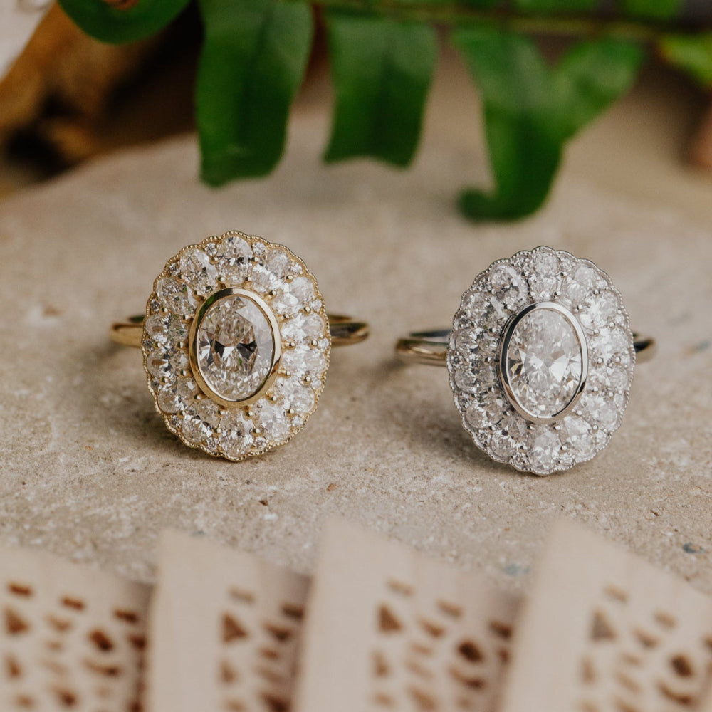 Two oval diamond halo rings in white and yellow gold on a textured surface with a leafy branch in the background