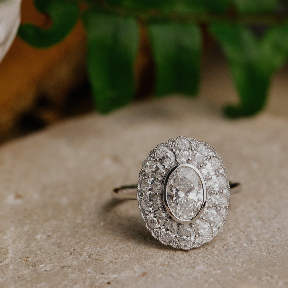 Oval diamond halo ring with a floral design on a textured surface with greenery in the background