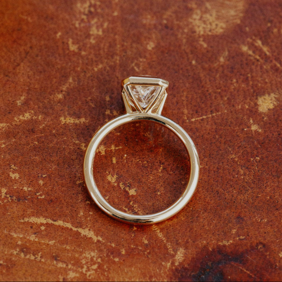 Gold Bailey bezel set engagement ring with a radiant cut lab grown diamond on a textured brown surface.