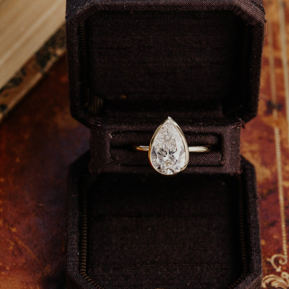 The Bailey pear-shaped diamond ring set in a bezel in a black velvet box on a textured surface.
