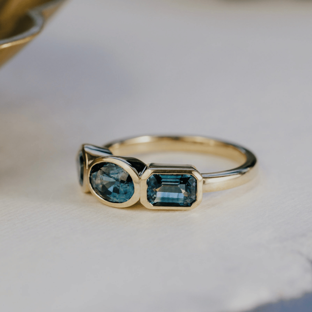 A gold ring featuring three blue sapphire gemstones in different shapes, resting on a light textured surface.