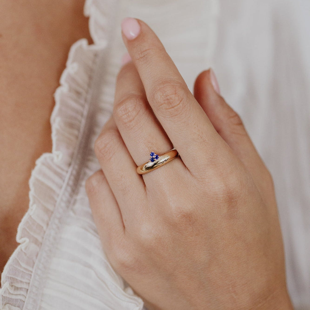 Hand wearing a gold ring with a small blue gemstone, against a blurred white fabric background.
