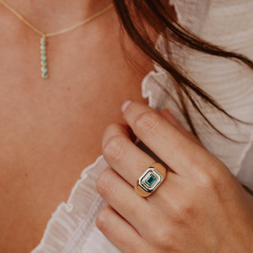 Close-up of a hand wearing a gold ring with a green emerald gemstone, with a blurred background.