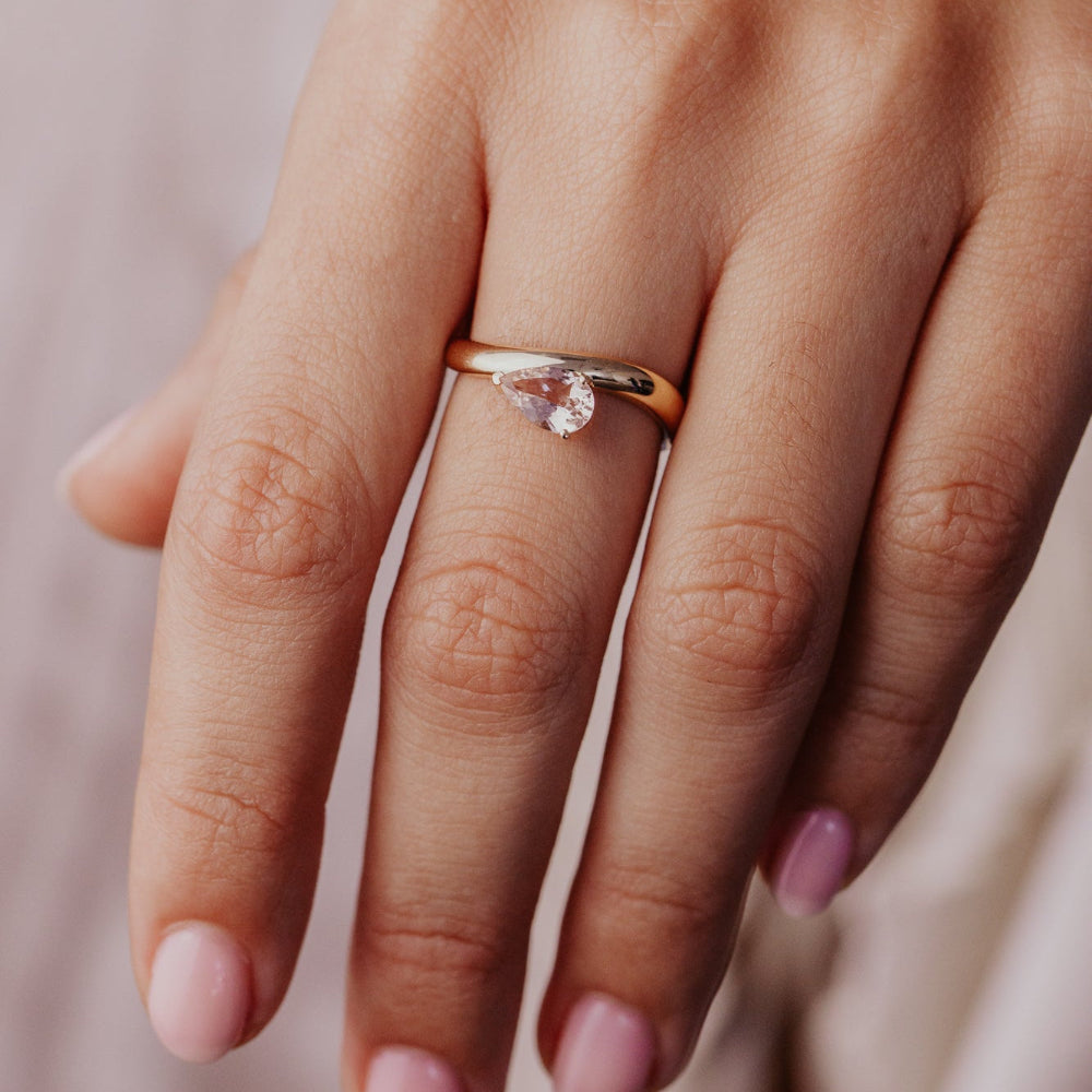 Close-up of a hand wearing a gold ring with a diamond on a soft pink background