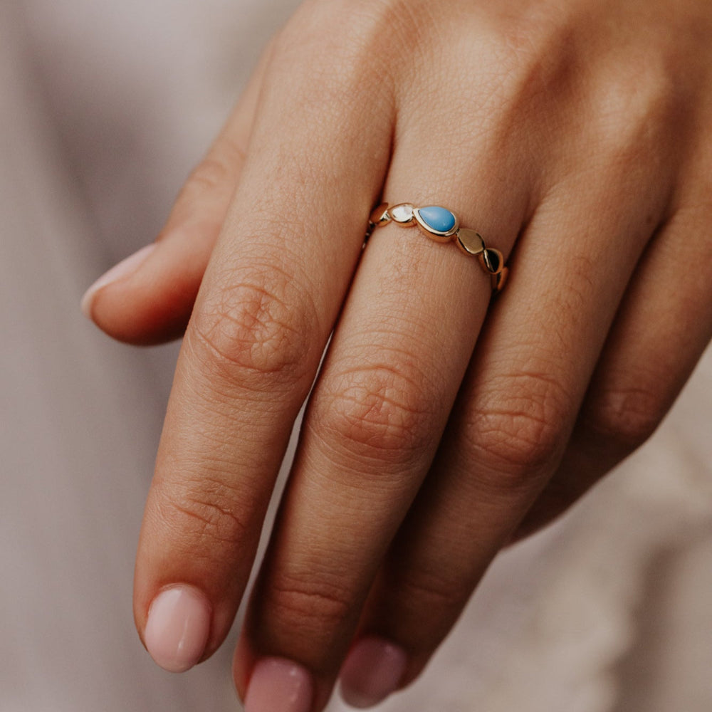 Close-up of a hand wearing a gold ring with a blue turquoise gemstone on a neutral background