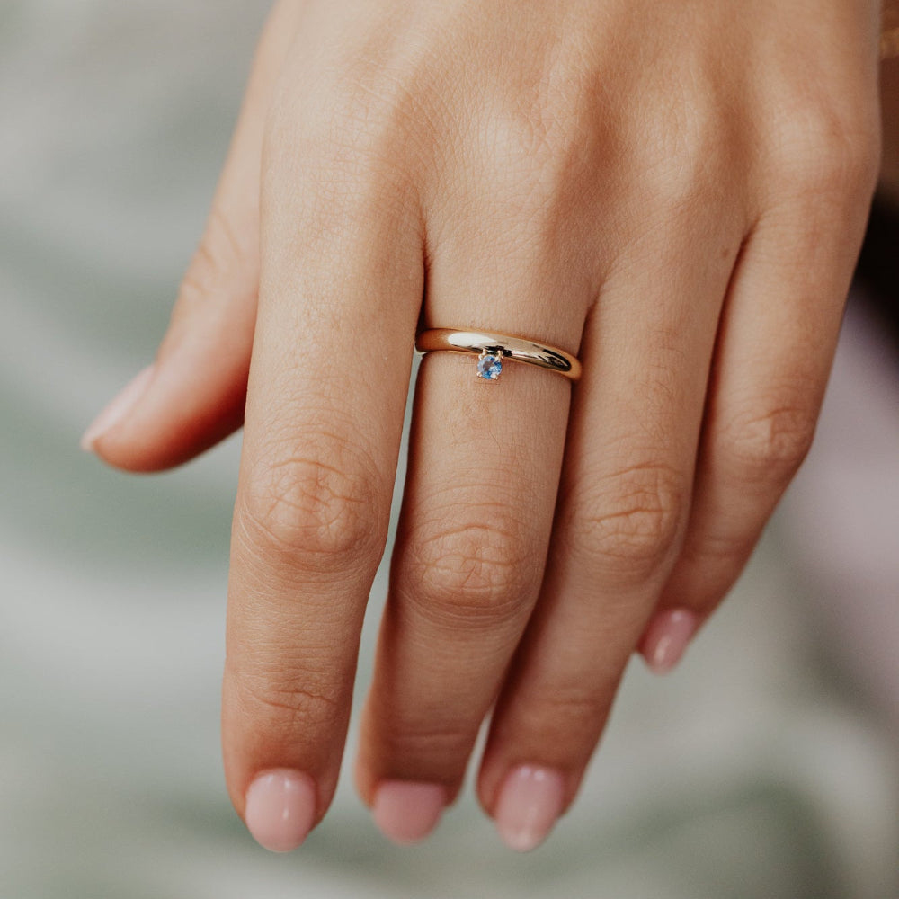 Close-up of a hand wearing a gold ring with a blue gemstone on a blurred background