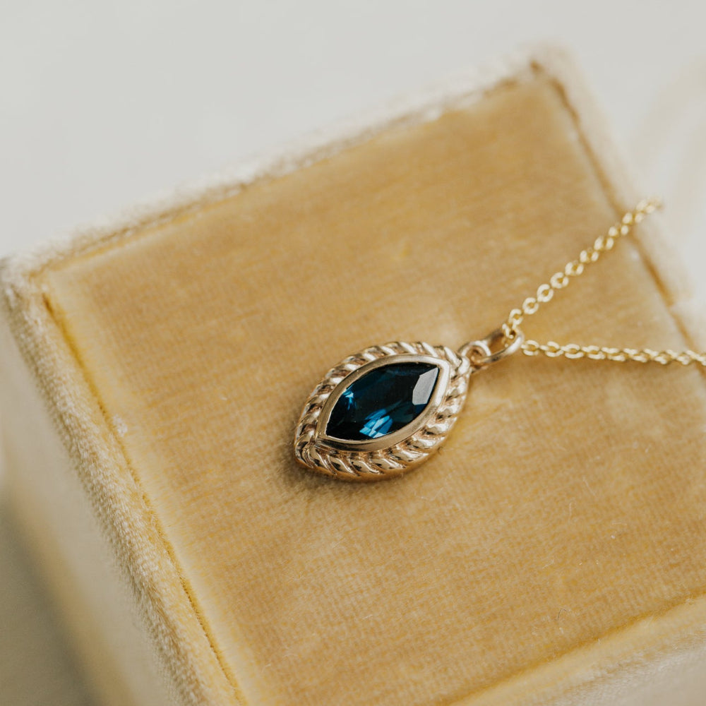 Gold necklace with a London blue topaz gemstone pendant on a beige velvet surface