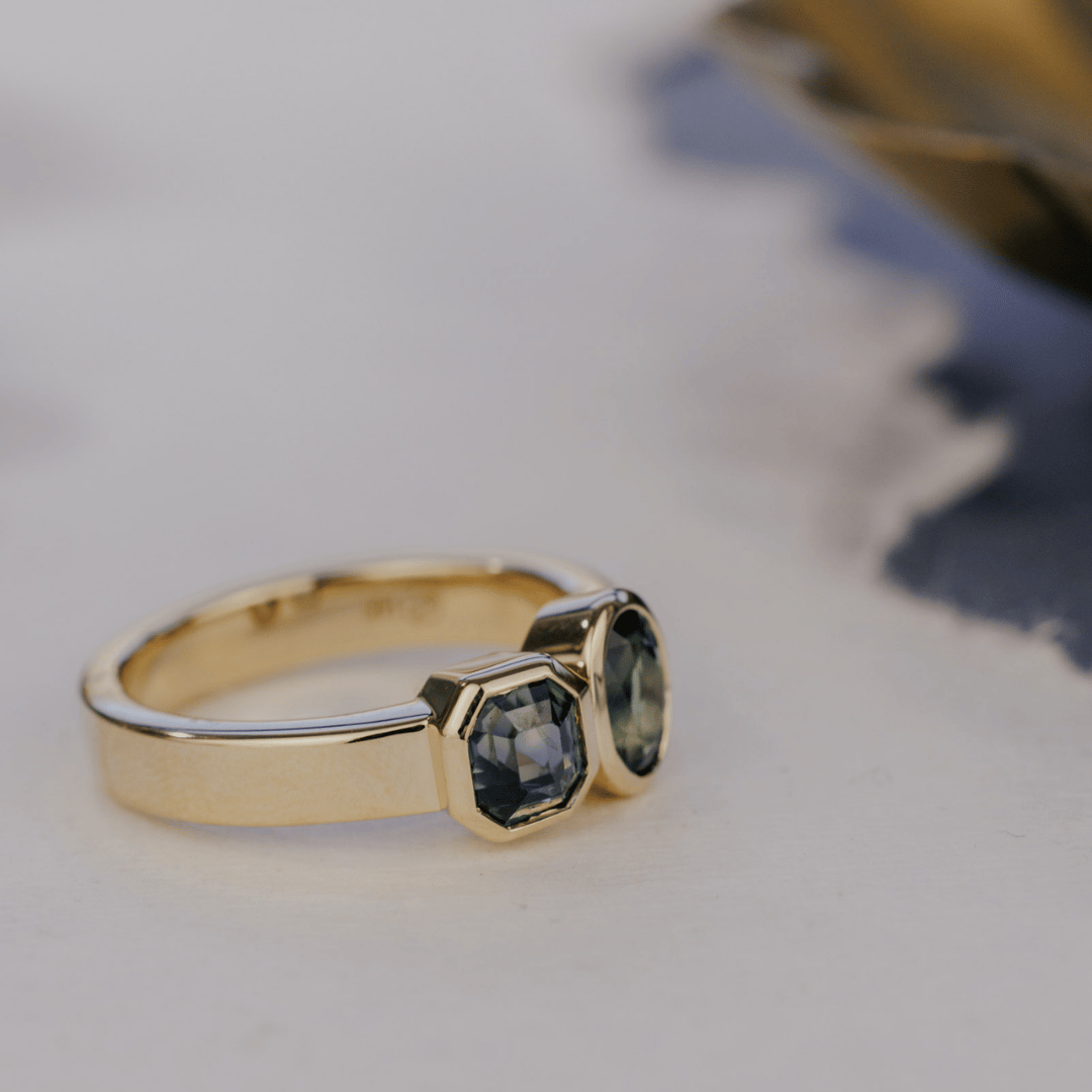 A 14k gold, two-stone ring with bezel-set Asscher and oval blue/teal sapphires in yellow gold. Set on a white surface with a soft-focus background.
