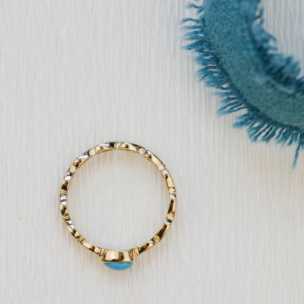 Gold ring with a blue turquoise gemstone on a white surface with a teal woven object.