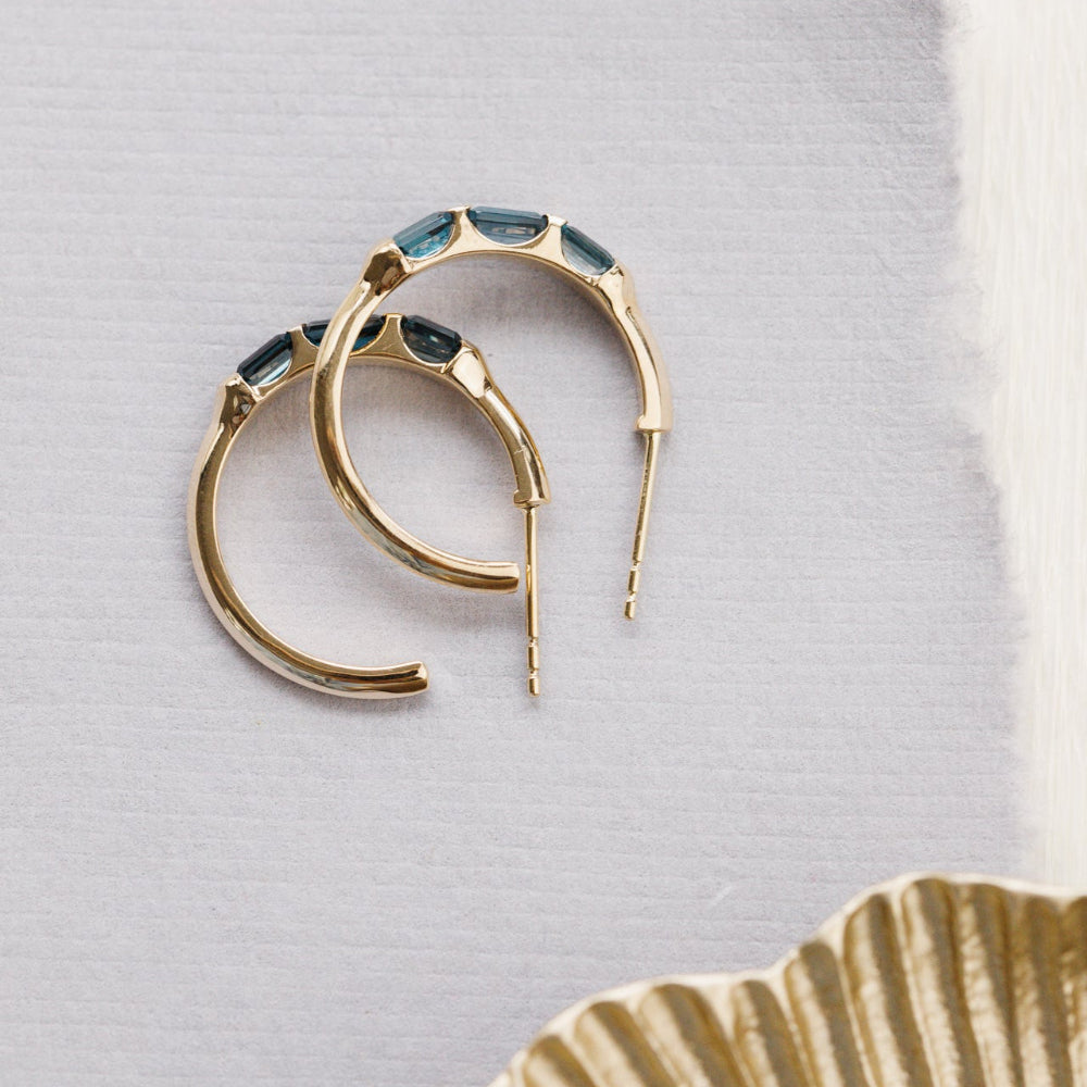 Gold hoop earrings with blue topaz gemstones on a light gray background