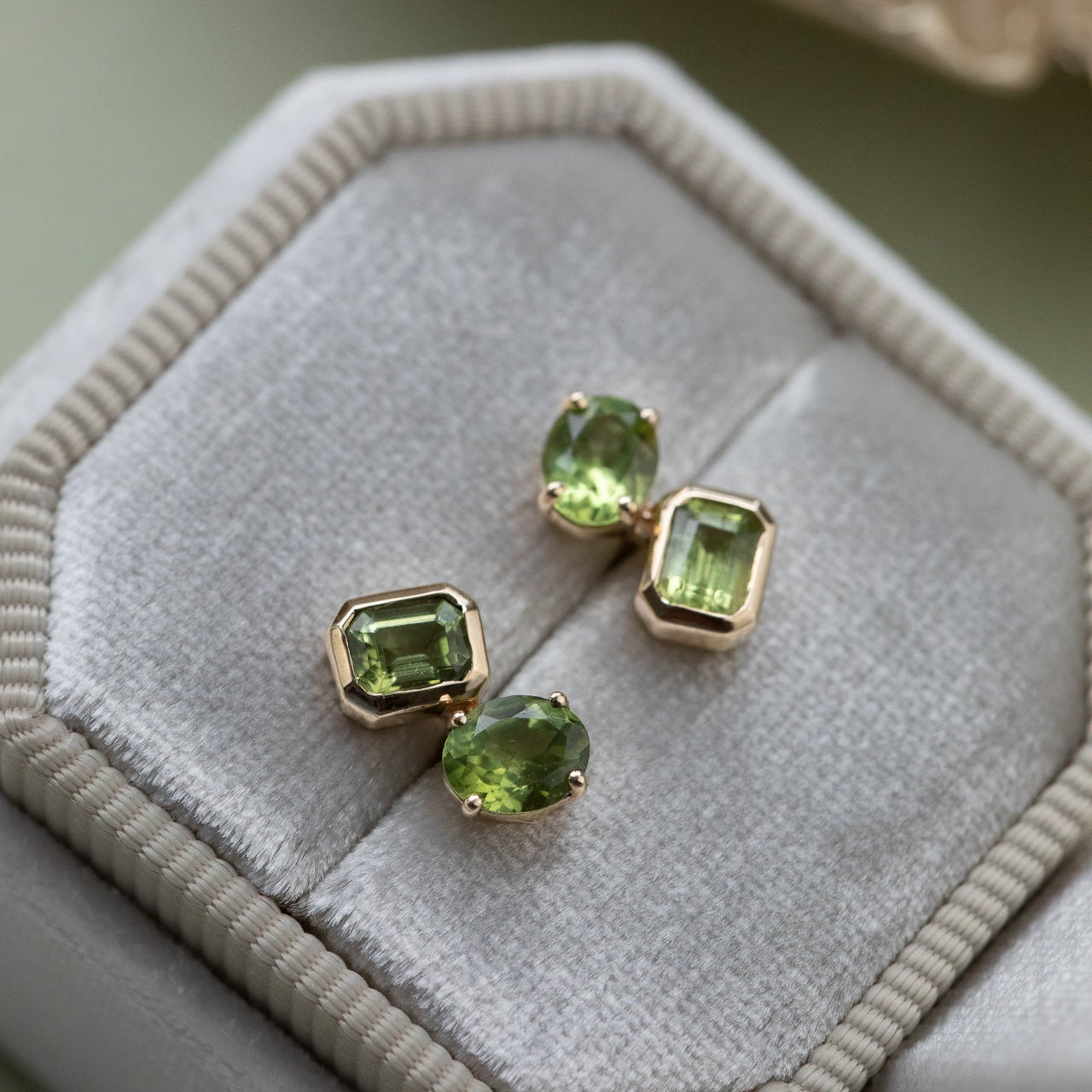 Green gemstone earrings on a hexagonal gray velvet cushion with a beige shell in the background.