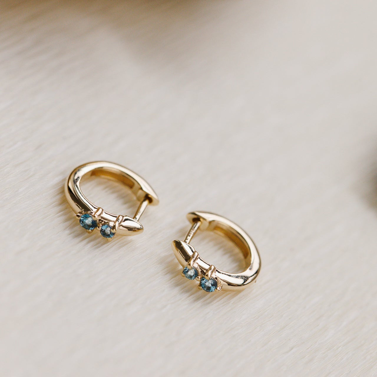 Gold hoop earrings with blue topaz gemstones on a beige surface