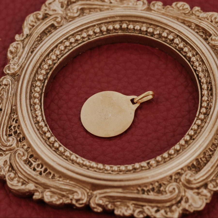 The Medium 18k Yellow Gold Vintage Love Token with Knife Edge and Rounded Bail by A. Augis, showing the smooth reverse side of the round 18k yellow gold vintage love token, displayed within an ornate gold oval frame on a deep red surface alongside a vintage paper prop.