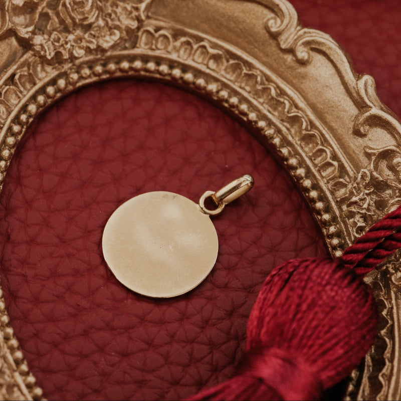 The Medium 18k Yellow Gold Vintage Love Token with Ruby Baguettes by A. Augis, showing the smooth reverse side of the round 18k yellow gold pendant, displayed within an ornate gold oval frame on a deep red surface alongside a burgundy tassel.