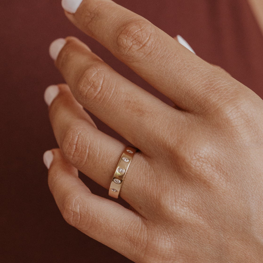 Close-up of a hand wearing a gold wedding band with marquise diamonds on a brown background