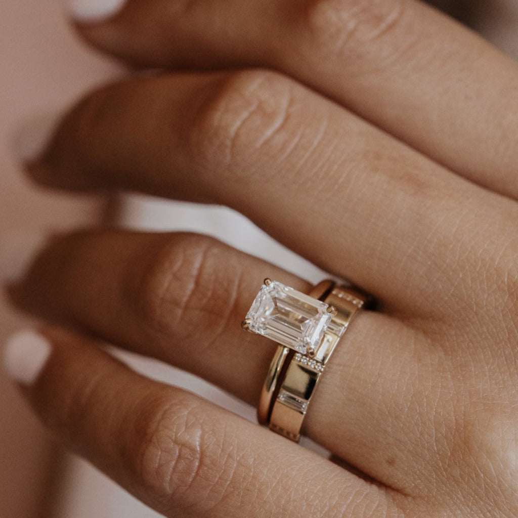 Close-up of a hand wearing a gold emerald cut engagement ring with a gold diamond wedding band on a blurred background