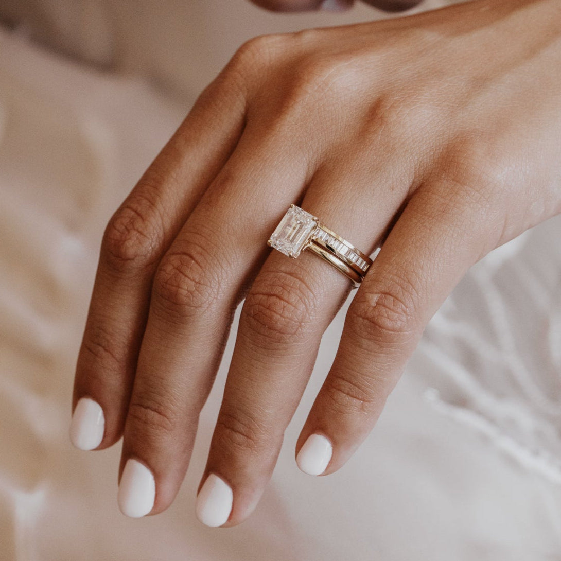 Close-up of a hand wearing a gold emerald cut engagement ring with a diamond wedding band on a soft background