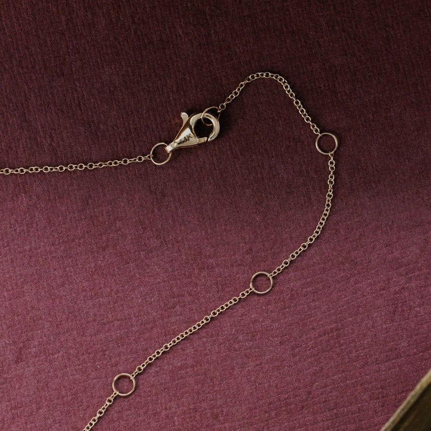 A close-up of a delicate gold chain necklace with a lobster clasp and three small circular ring details, laid on a textured dark burgundy surface.