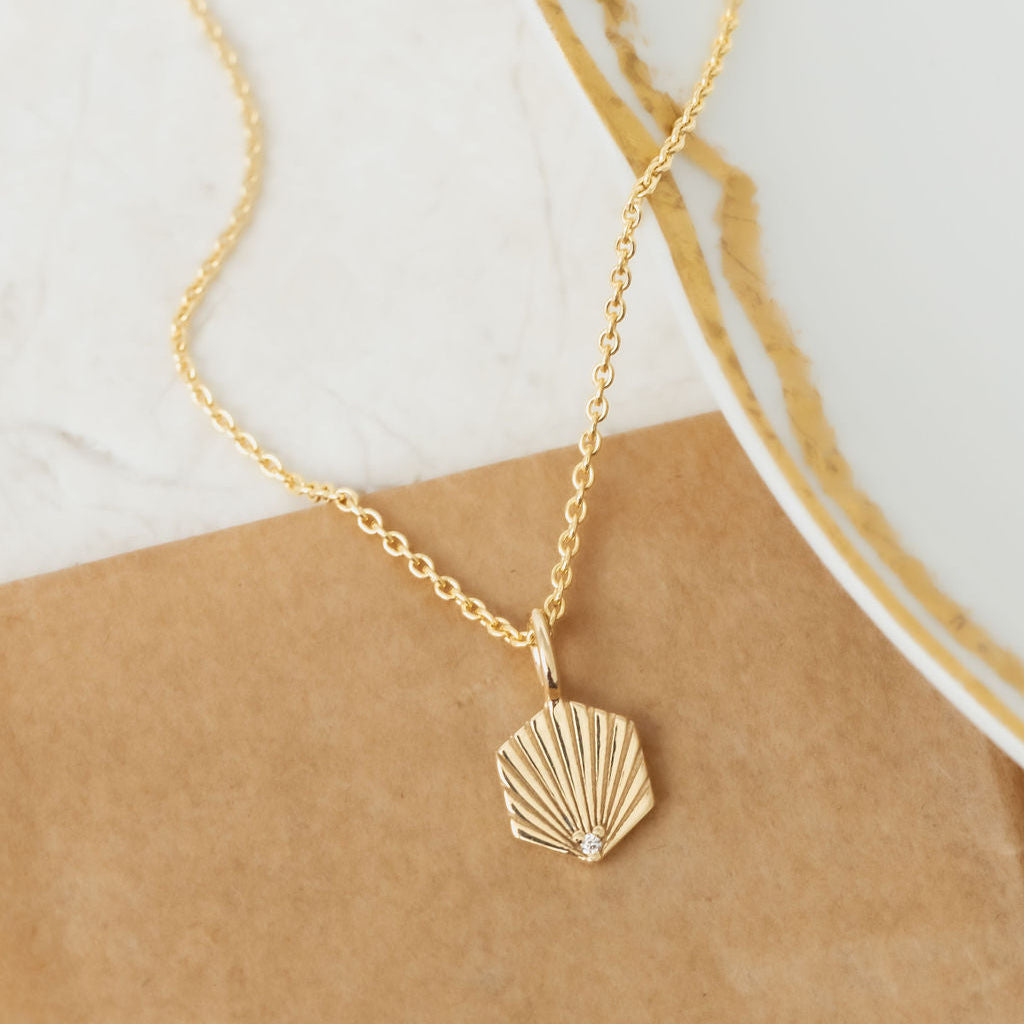 A dainty gold necklace with a hexagon shell shaped pendant and a round pave diamond at the center, displayed on a white surface.