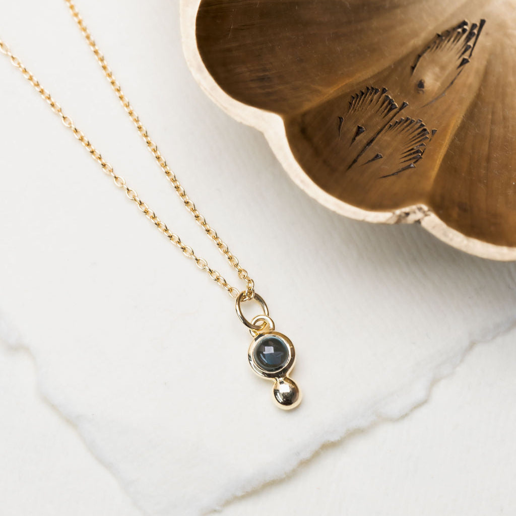 A round blue topaz gemstone necklace with a metal ball accent, displayed on a white dish with a gold-colored metal chain.