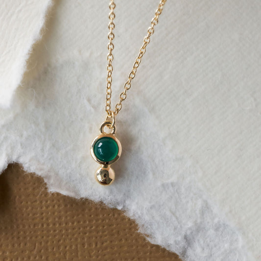 A gold necklace with a round green agate set in a bezel and a metal ball accent, displayed against a white and brown background.