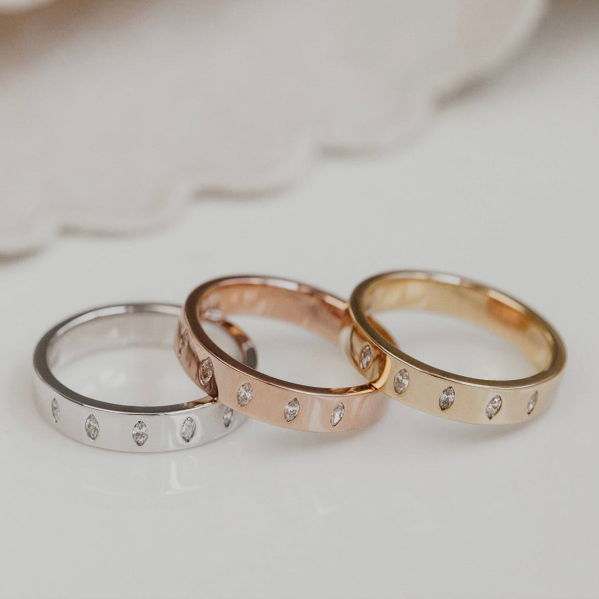 Three wedding band in yellow gold, rose gold, and white gold on a white surface with a blurred background.