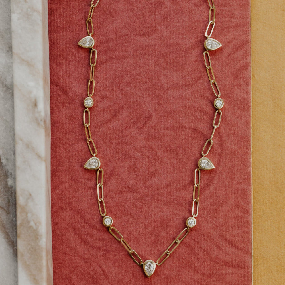 Gold chain necklace with pear-shaped diamond pendants on a textured red surface.