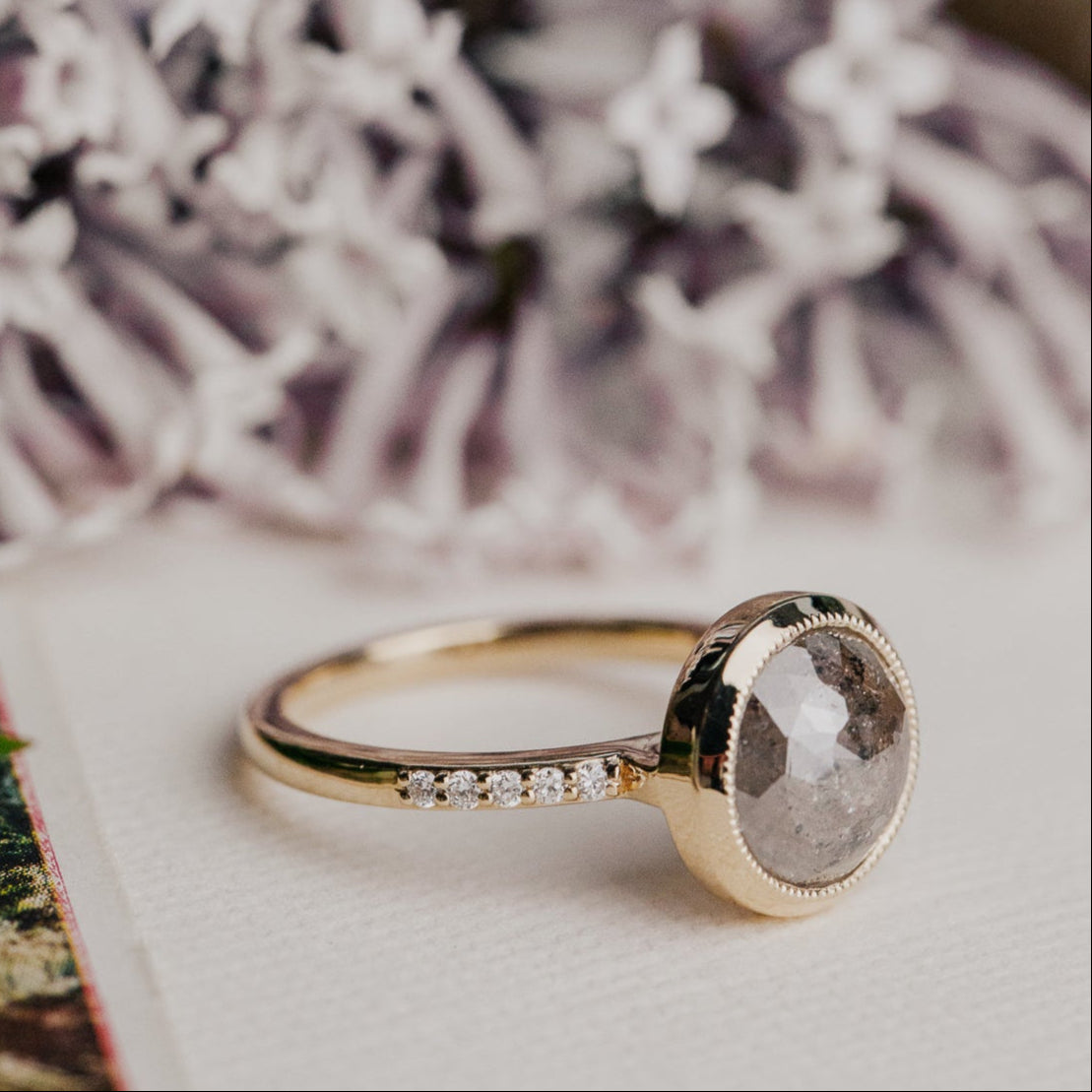 14k gold ring featuring a round grey rustic diamond set in a milgrain bezel with a .09 cttw pave diamond band.