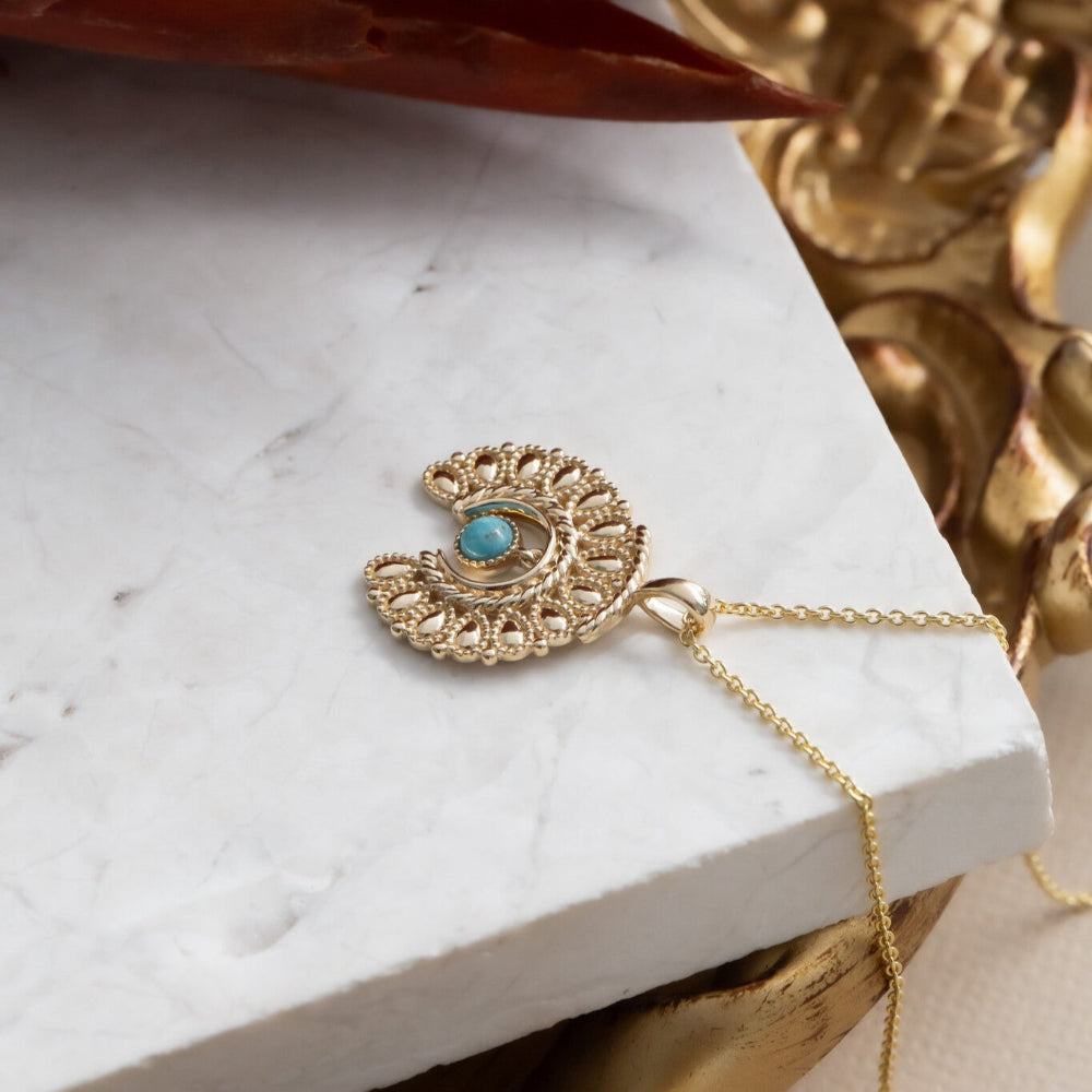 Gold necklace with a turquoise pendant on a marble surface with decorative elements.