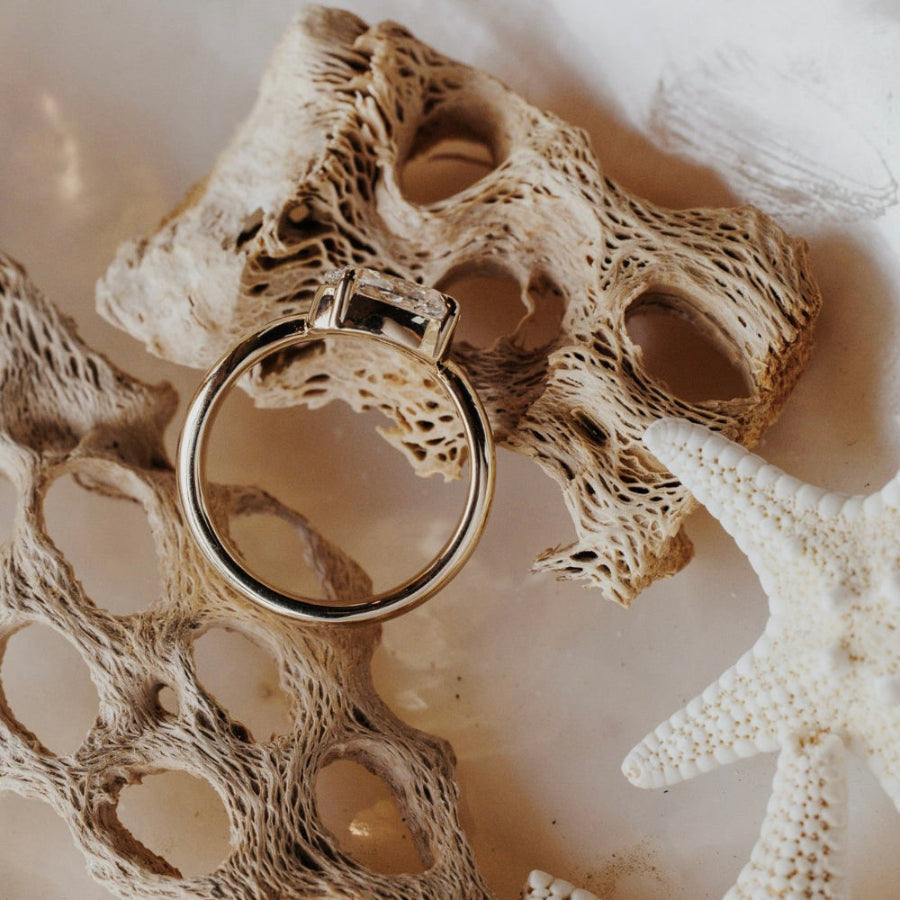 The Capri Oval Ring in 14k yellow gold featuring a lab-grown oval diamond prong set on a double wavy band, shown from above on coral and starfish.
