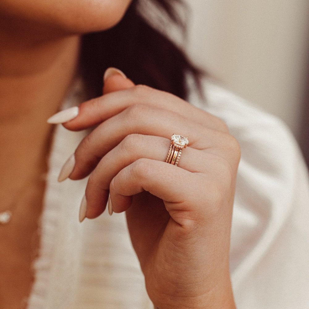 The Claire Oval Ring in 14k rose gold with a lab grown diamond oval center stone, worn stacked with multiple bands on a model's hand against a white fabric background.