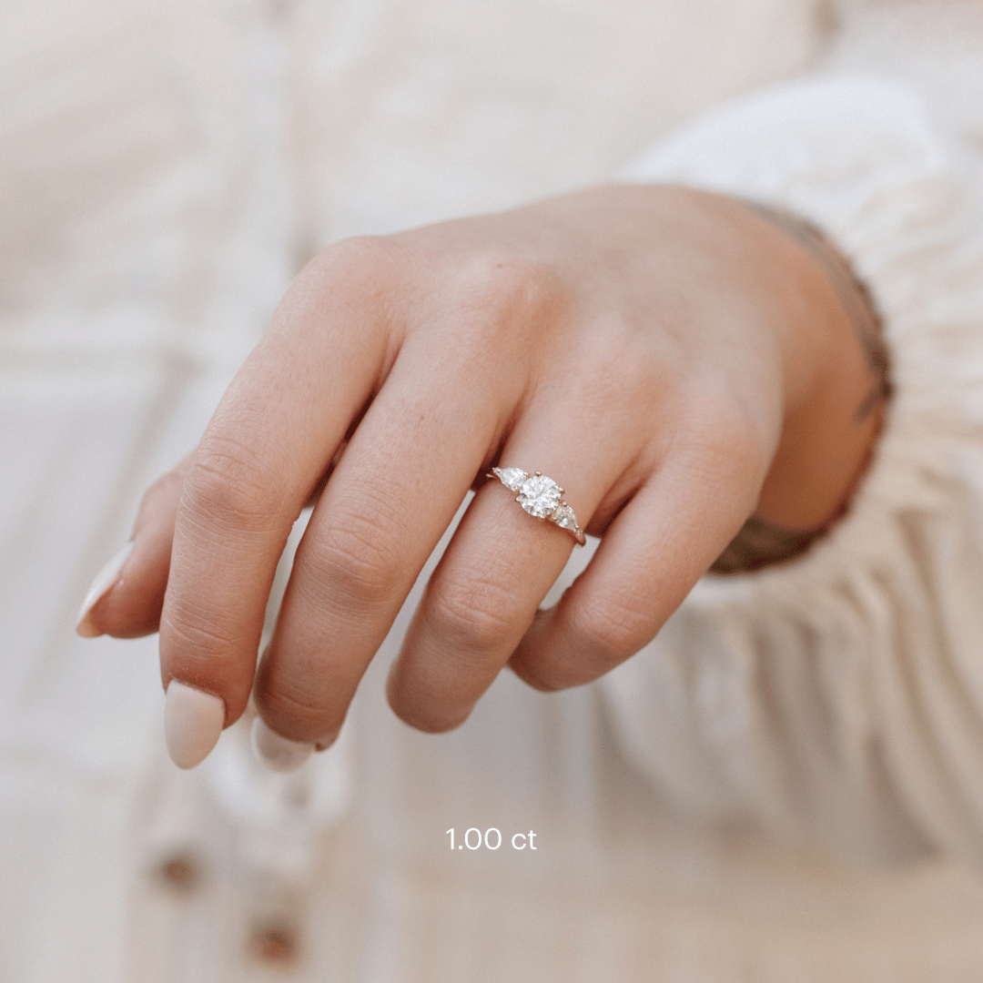 The Toula Round Ring in 14k yellow gold featuring a lab grown diamond round center stone with pear and round natural diamond side stones, worn on a model's outstretched hand against a cream textured background.