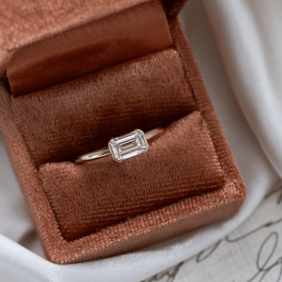 Austen diamond ring in an open brown velvet box on a white surface with a piece of paper.