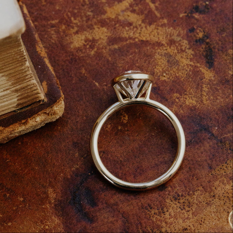 Gold Bailey oval diamond engagement ring on a textured brown surface.