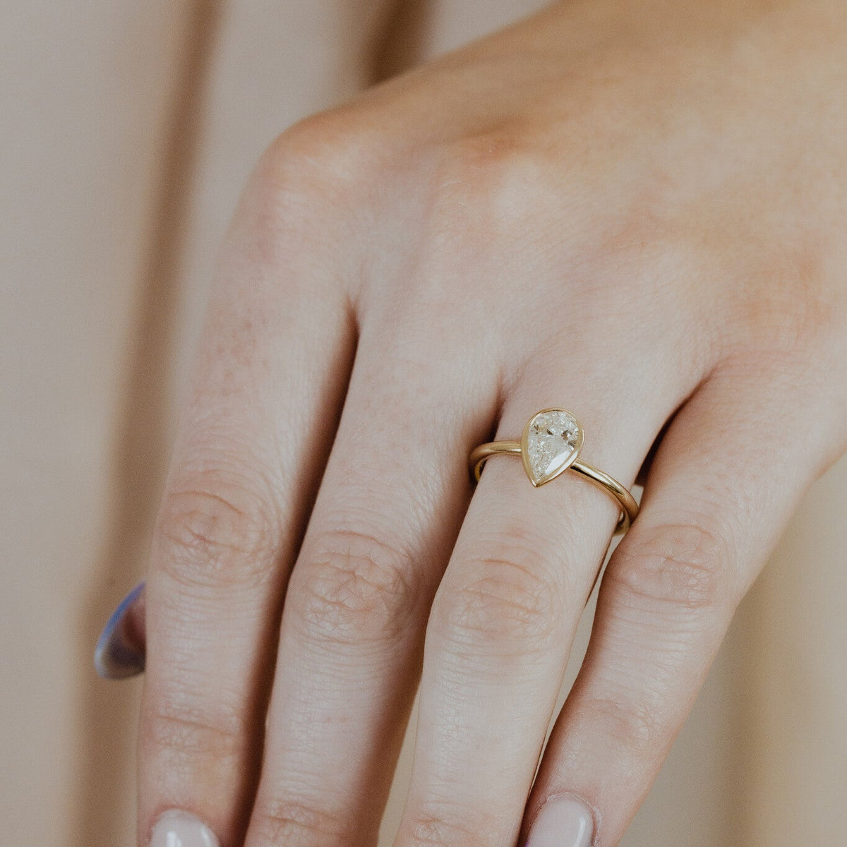Hand wearing a gold Bailey ring with a pear-shaped diamond on a neutral background.