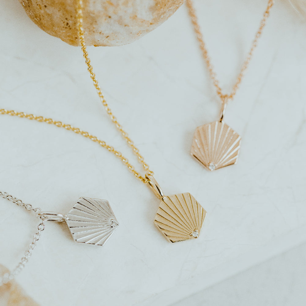 A dainty gold necklace with a hexagon shaped pendant and a round pave diamond at the center, displayed on a white surface.