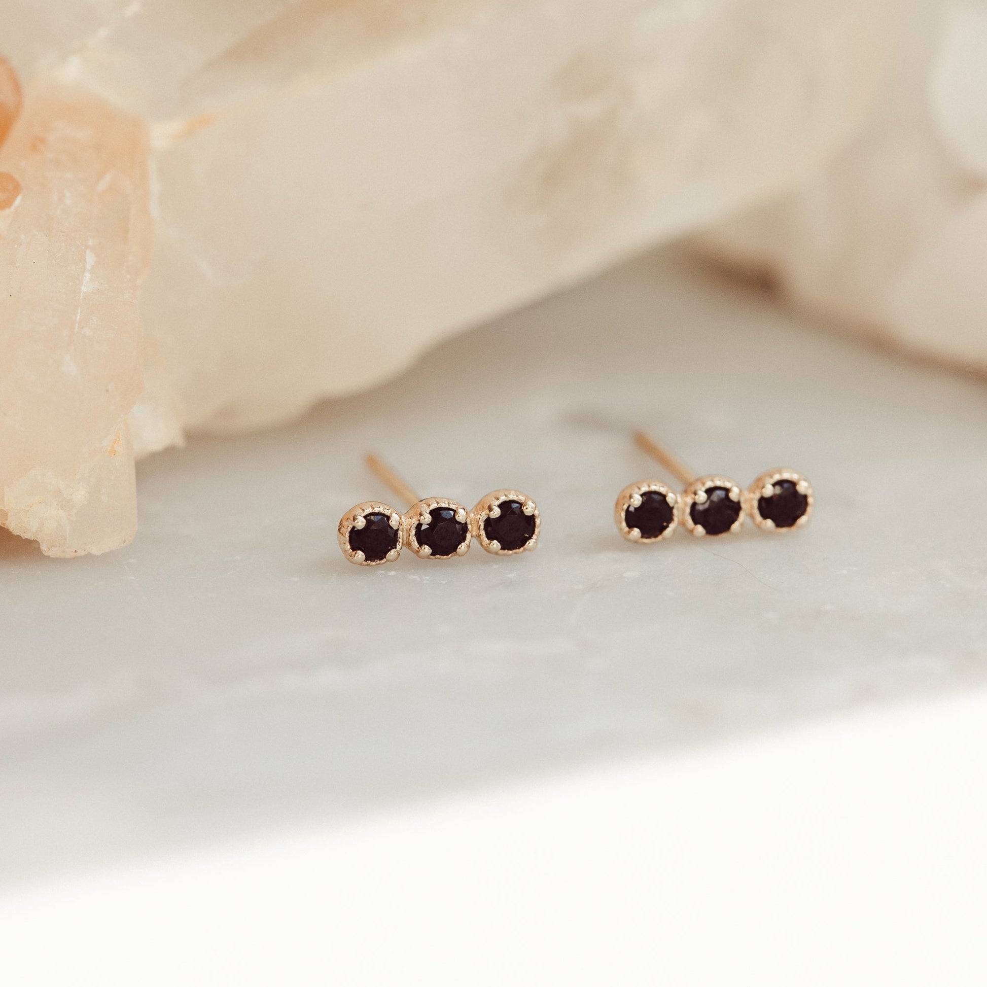 A pair of 14k gold stud earrings featuring three round black spinels set in prongs, displayed against a light-colored crystal background.