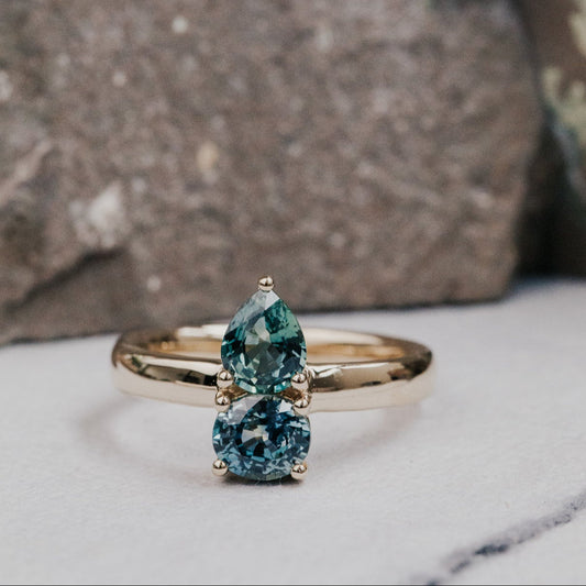 Gold ring with two green Montana sapphire gemstones on a marble surface with a stone background