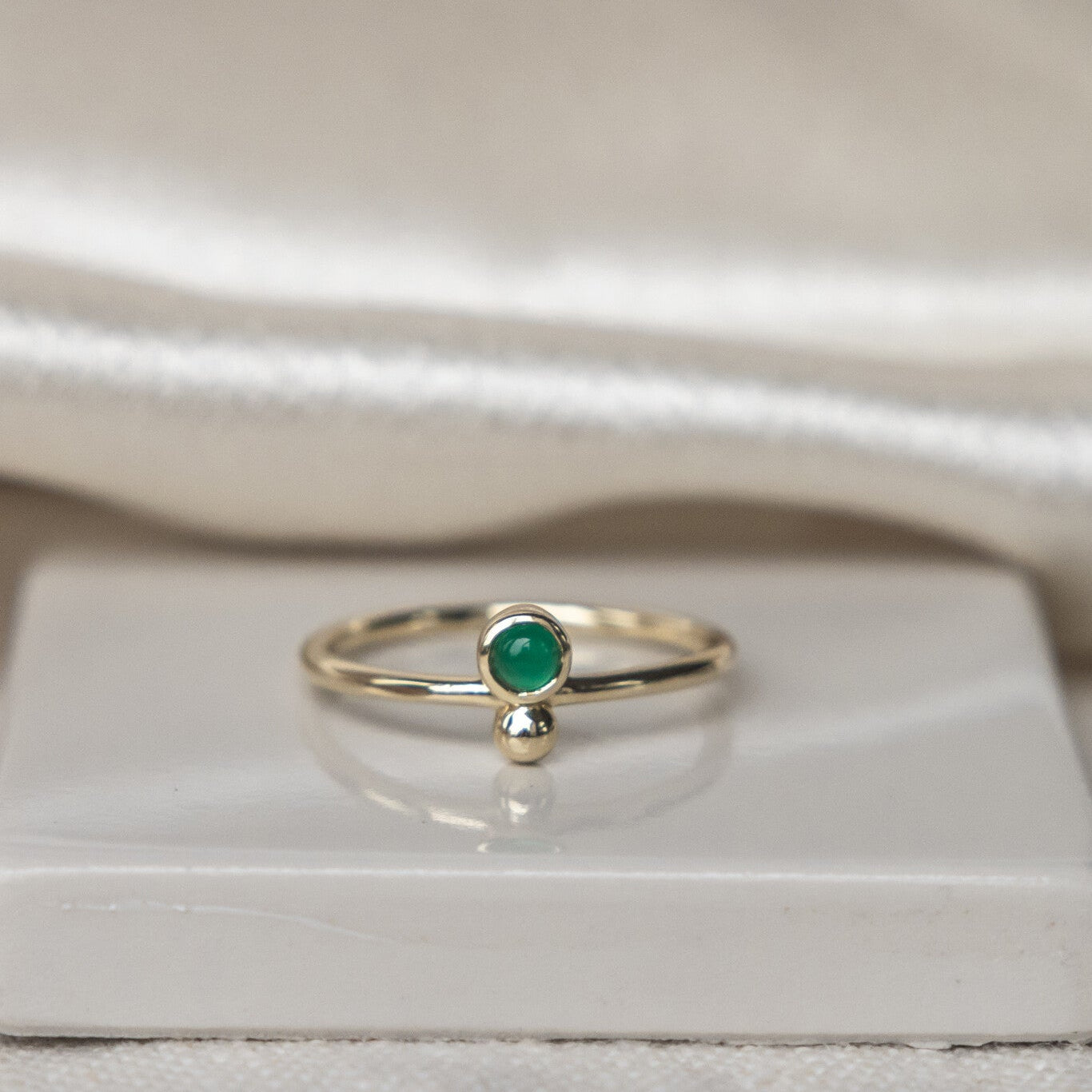 A 14k gold ring with a round green agate center stone set in a high polish bezel, accompanied by a metal ball accent on a thin band, displayed on a white surface.