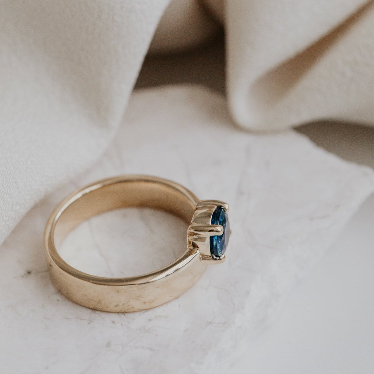 Gold Sezanne ring with a blue Montana sapphire gemstone on a soft beige fabric background.