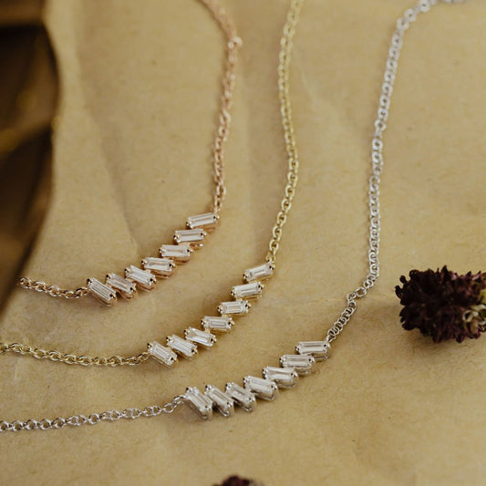 A curved bar necklace with baguette diamonds set in gold, displayed on a beige background with some floral elements.