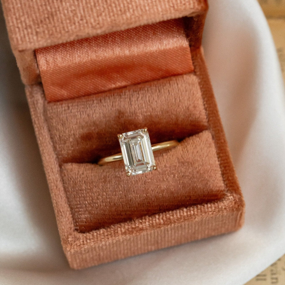 An emerald cut diamond solitaire ring in a rose gold band, presented in a brown velvet box.