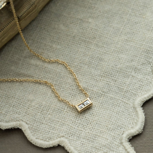 A gold necklace with two baguettes set side by side in a high polish bezel, displayed on a beige fabric surface.