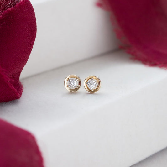A pair of 14k yellow gold stud earrings with .11 ct round diamonds set in an organic metal halo, displayed on a white surface with a red textured background.