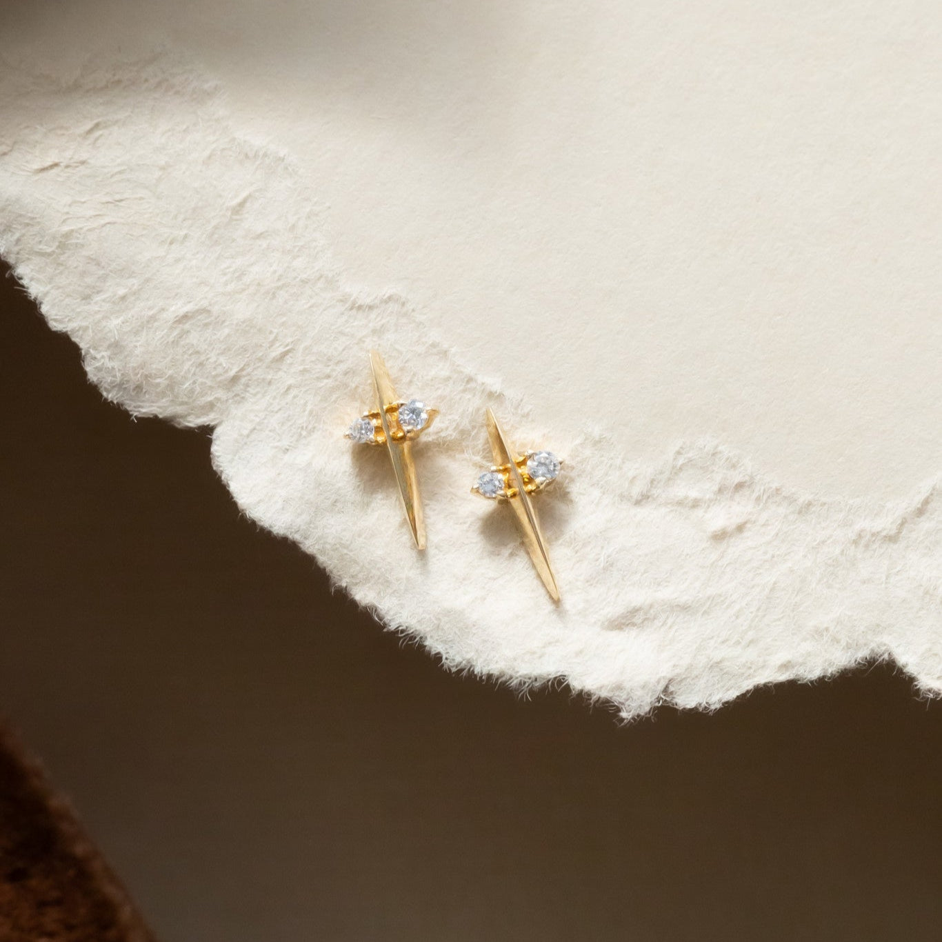 The image shows a close-up view of a pair of 14k yellow gold stud earrings with round blue diamonds set in prongs on a tapered bar, displayed on a soft surface.